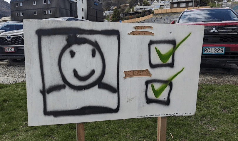 A wooden sign displays a spray-painted smiley face next to two checkboxes, both marked with green checkmarks. It stands outdoors on grass, with parked cars and buildings in the background.