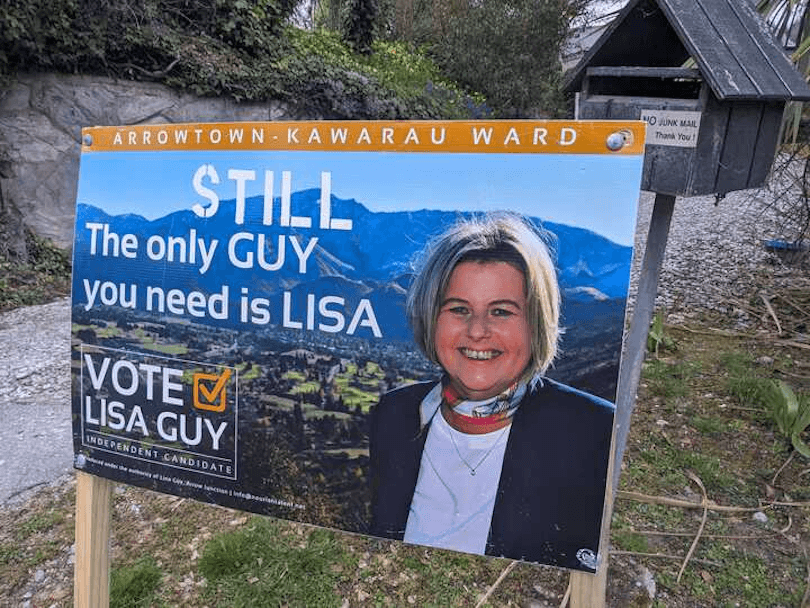 A campaign sign for Lisa Guy in Arrowtown-Kawarau Ward reads, “Still the only GUY you need is LISA.” It features Lisa Guy’s portrait with mountains in the background and encourages voting for her.