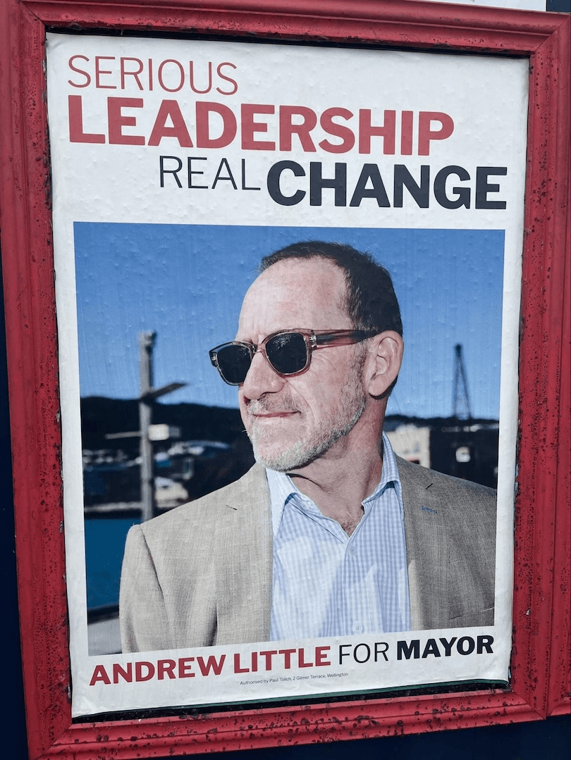 A campaign poster featuring a man in sunglasses and a beige jacket. The text reads: "Serious Leadership, Real Change. Andrew Little for Mayor." The background shows an outdoor scene with blue sky.