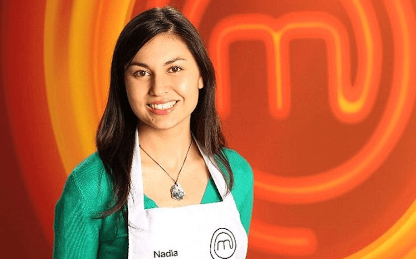 A young Nadia Lim wears a green top and white MasterChef apron in front of the neon orange MasterChef logo