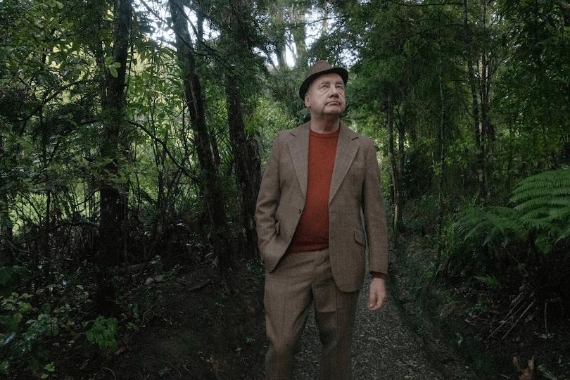 A man wearing a brown suit, matching hat, and orange sweater walks along a wooded forest path, surrounded by green trees and foliage.