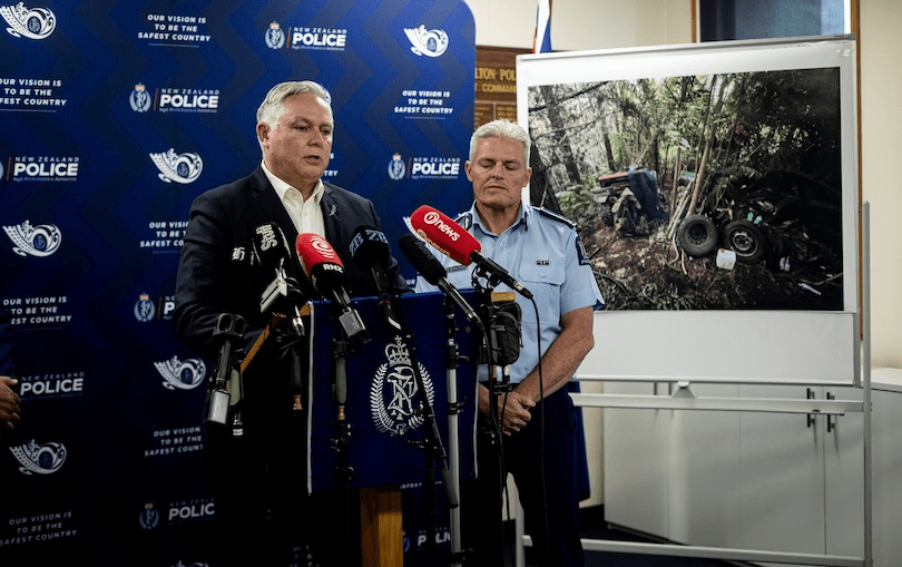Two New Zealand police officers stand at a podium with microphones during a press conference, with a large photo of a crashed vehicle in a forest displayed on an easel beside them.