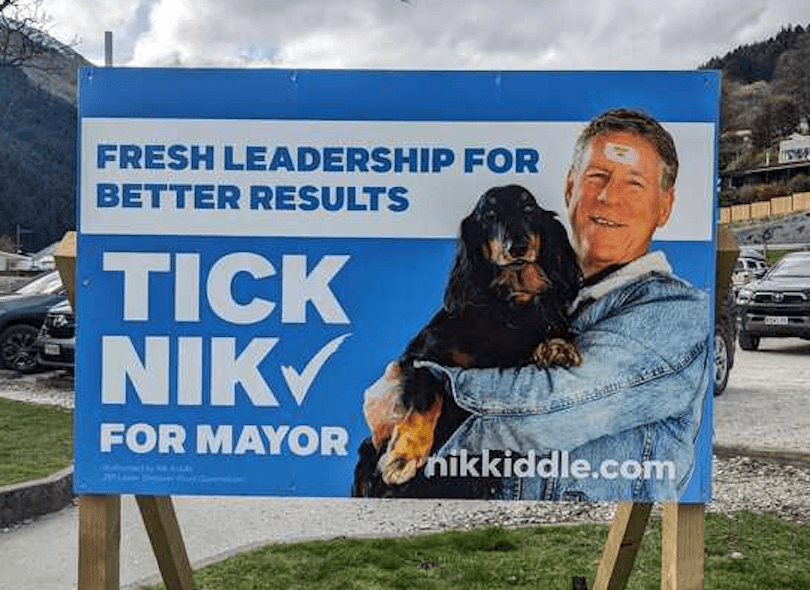 A campaign billboard features a man with a bandage on his forehead holding a black and brown dog. The sign reads, “Fresh leadership for better results. Tick Nik for Mayor. nikkiddle.com.”.