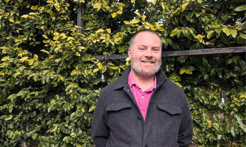 a middle aged man, bald, smiling, pink shirt black jacket, with a bush behind him