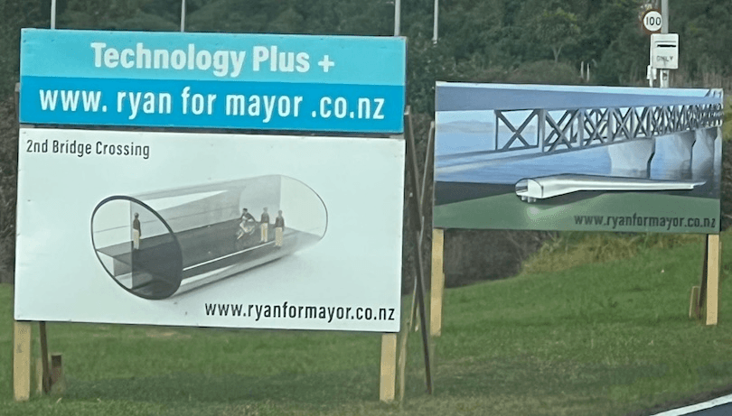 Two campaign signs read "Technology Plus + www.ryanformayor.co.nz." One shows a tube-like bridge with people inside labeled "2nd Bridge Crossing," the other shows a bridge over water. Grass and trees are in the background.