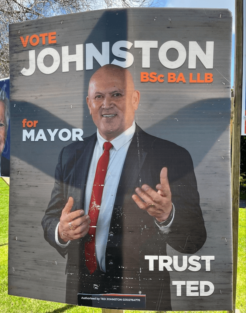 Campaign poster showing a bald man in a suit and red tie, smiling and gesturing with his hands. Text reads: “VOTE JOHNSTON BSc BA LLB for MAYOR. TRUST TED.” The poster is displayed outdoors on a sunny day.