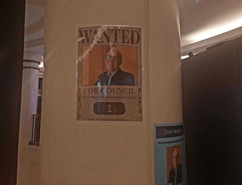 A poster on a beige pillar features an older man in glasses and a suit with the large text "WANTED for Council" above his photo. Another smaller poster with his photo is partially visible on the right.