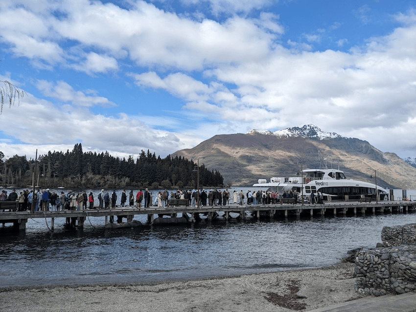 a line of people on a wharf with a boat infront of them and some mountains and a lake behind them