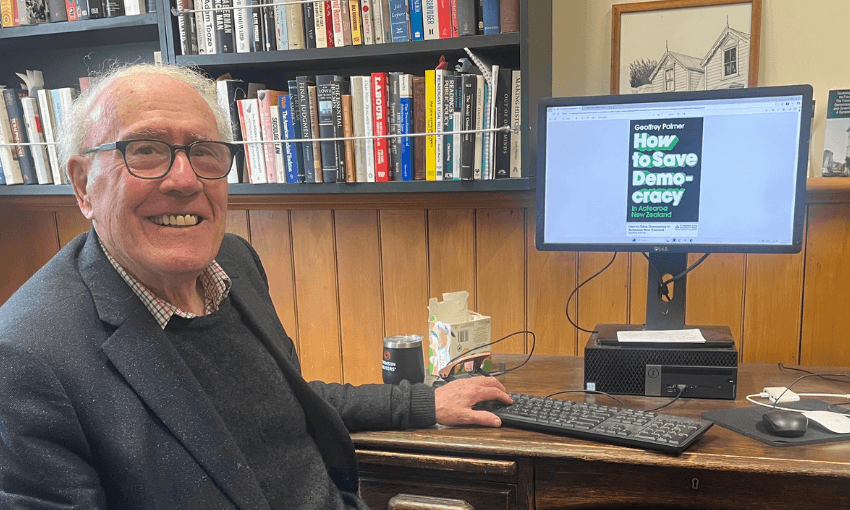 Geoffrey Palmer sits and smiles on front of his desk, with his compute displaying the cover of his book How to Save Democracy.