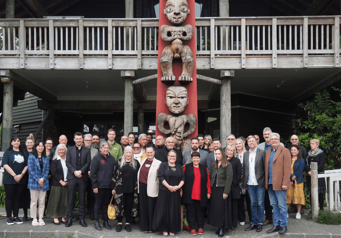A large group of people stand in front of a carved wooden totem pole and a traditional wooden building, posing for a group photo. The scene is outdoors, with greenery visible on the right.