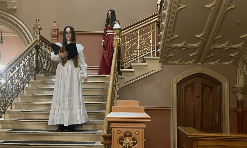 Two women wearing Victorian gowns stand on the main staircase in parliament's library area.