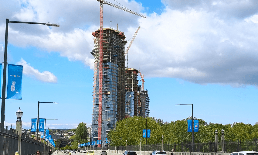 Two high-rise buildings under construction with cranes on top are viewed from a bridge lined with blue banners, under a partly cloudy sky. Cars and people are visible on the bridge in the foreground.