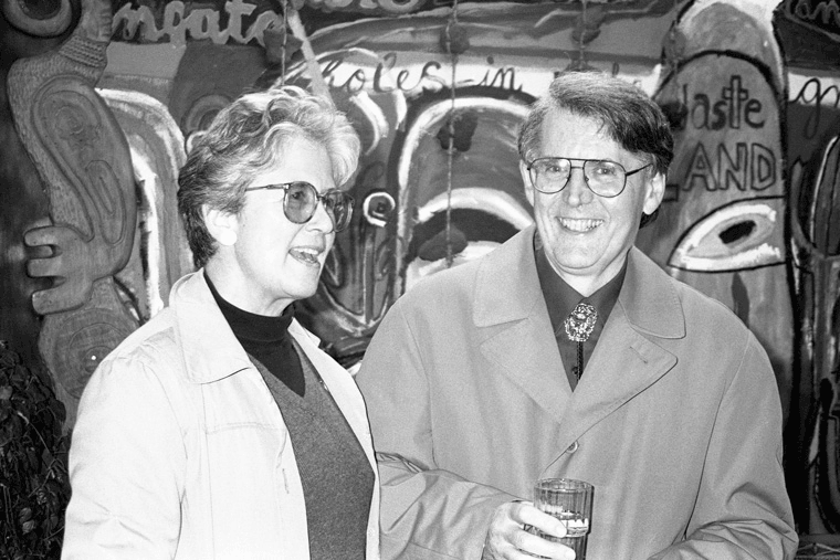 A black-and-white photo of two smiling adults wearing glasses and coats, standing together. The man holds a drink. Colourful, graffiti-style art is visible on the wall behind them.