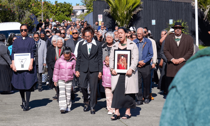 A large group of people attends a pōwhiri. Two women at the front carry framed photos, and two girls in pink jackets walk beside them. The crowd follows closely behind under sunny skies.