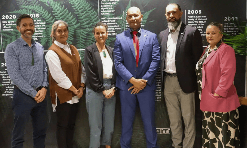 Six people stand in a row, dressed in business or smart casual attire, in front of a background featuring green ferns and text about historical events. They appear to be posing for a formal group photo.