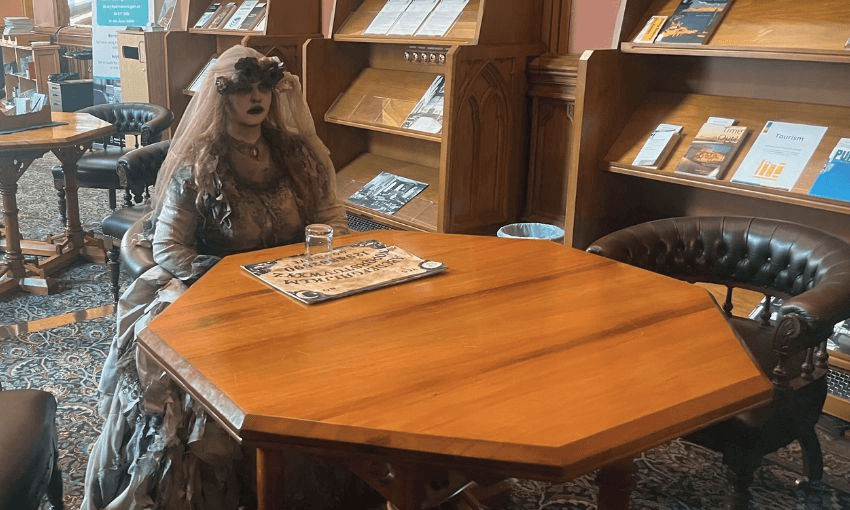 A woman wearing tattered Victorian garb sits at a table in parliament's library, with an ouija board in front of her.