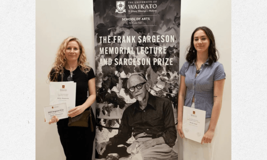 A photo of two women standing either side of a banner that shows a black and white photo of a man who was Frank Sargeson. The two women are both holding certificates. 