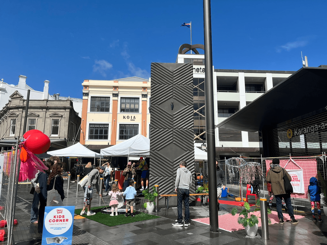 People, including families with children, enjoy activities at an outdoor event in a city square. There are tents, activity stations, and a sign for a kids' corner under a blue sky with modern and historic buildings in the background.