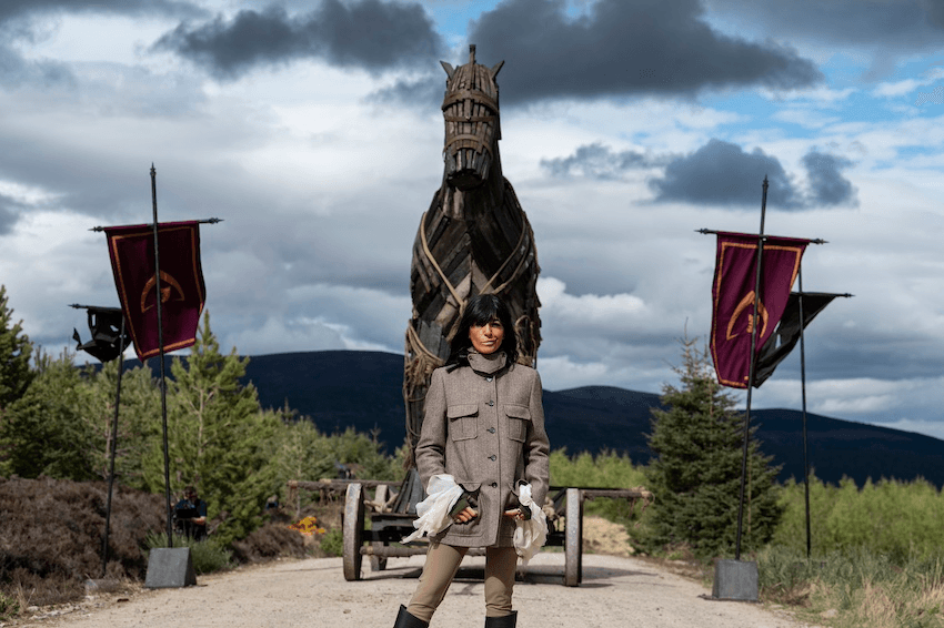 Claudia Winkleman stands on a gravel road with her hands in her pockets. Behindd her is a large Trojan horse and stormy skies