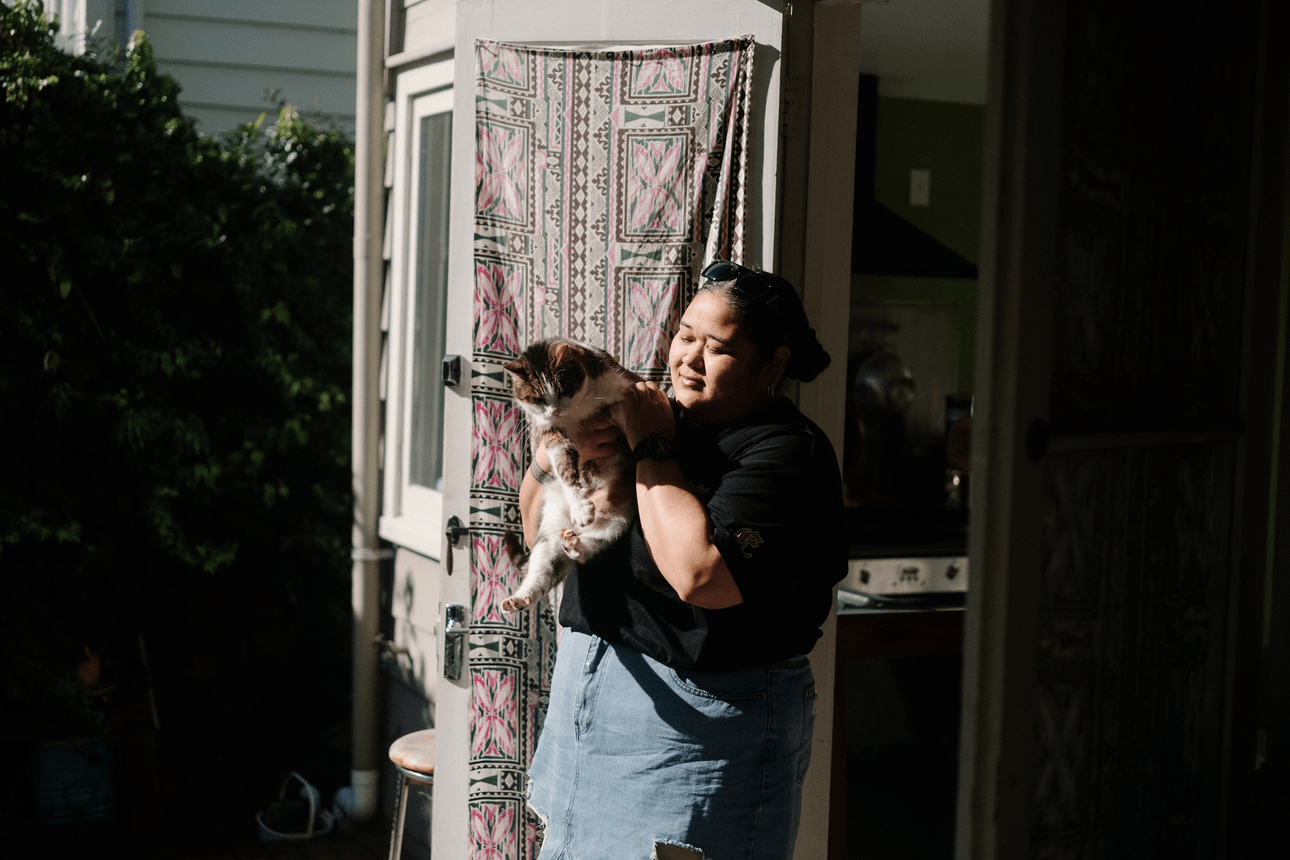 a woman in a black t-shirt and denim skirt stands in the sun holding a white, black and brown cat