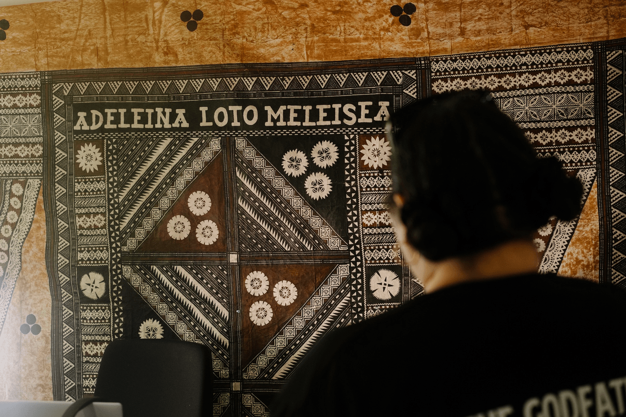 a tapa cloth in black and brown hangs on the wall with the profile subject's name printed on it. A woman with dark hair looks at it, facing away from the camera