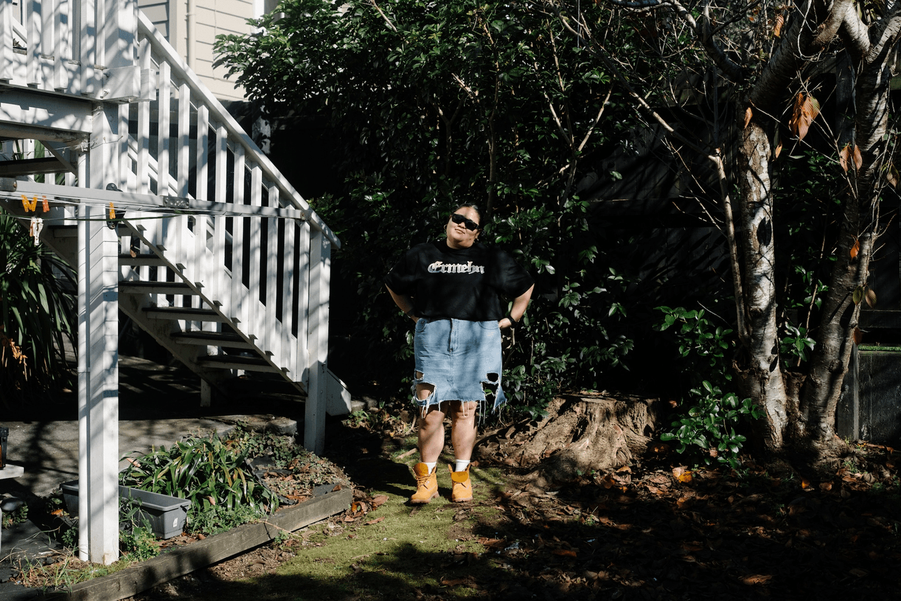 a young woman in a black t-shirt, denim skirt and Timbaland boots stands in her garden wearing dark sunglasses