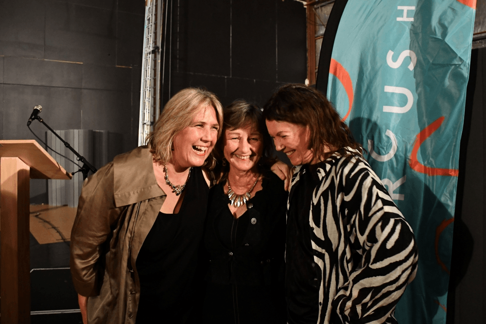 Three women stand together smiling and laughing in front of a teal banner and black backdrop. One wears a light jacket, one is in black, and one wears a patterned jacket. A wooden podium and microphone appear on the left.