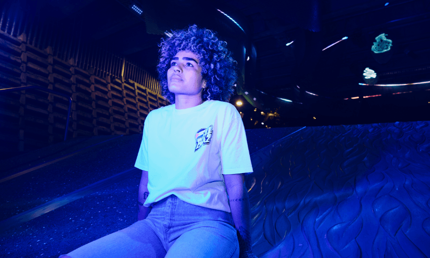 A person with curly hair sits on a metallic slide under blue and purple lights, looking upward. They wear a white t-shirt and jeans, with futuristic patterns and neon accents in the background.