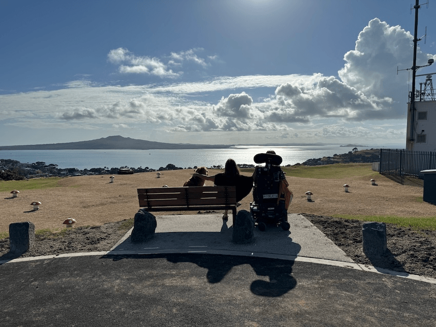a suny day looking over the cone of ranitoto, with a park bench with a person sitting in it and a power wheel chair next to her 