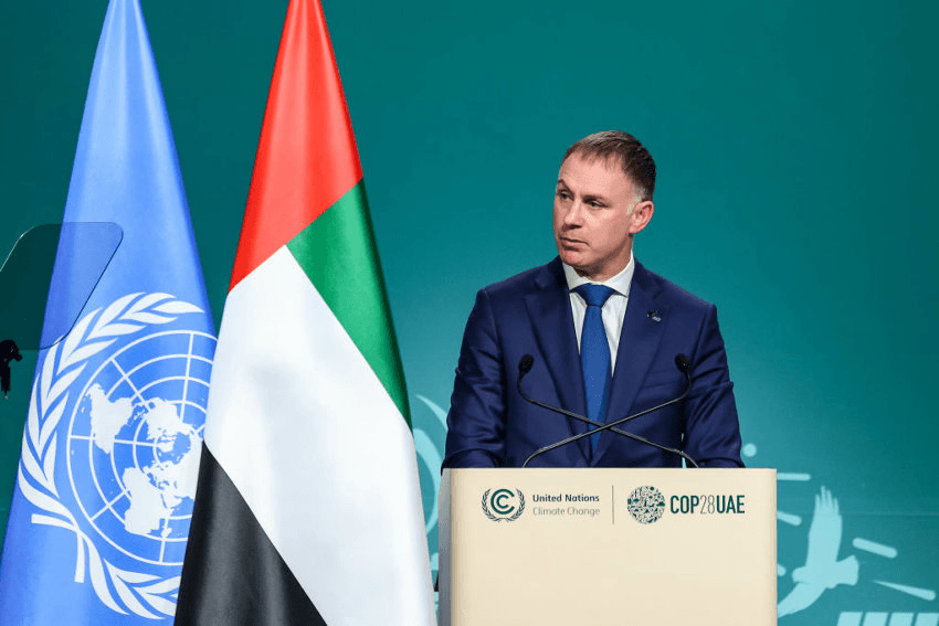 A man in a blue suit stands at a podium with the UN and UAE flags behind him, speaking at the United Nations Climate Change Conference (COP28 UAE) against a green backdrop.