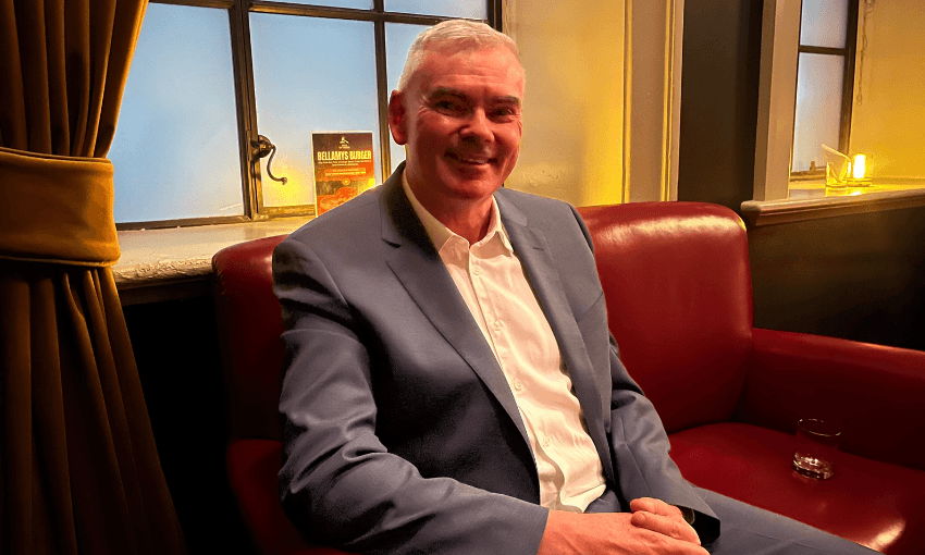Minister Scott Simpson, in a grey suit, sits on a red couch in a dark corner of Pint of Order.