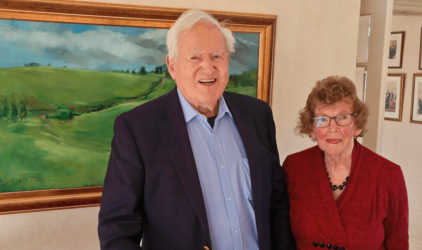 Jim and Joan Bolger in their Waikanae home in June 2025. In the background is a painting of their Te Kūiti farm. (Photo: Toby Manhire) 
