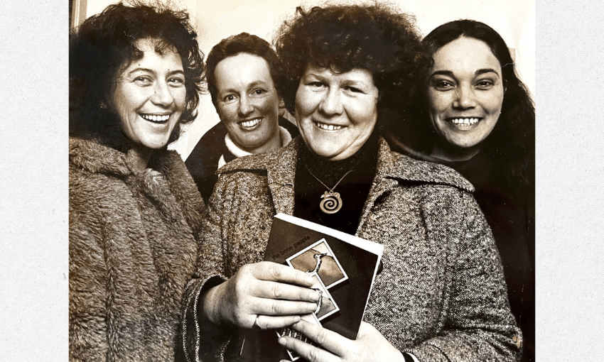 A black and white photograph of the writer Keri Hulme holding her novel the bone people and surrounded by three other women who make up the Spiral Collective who published the work. They are all smiling.