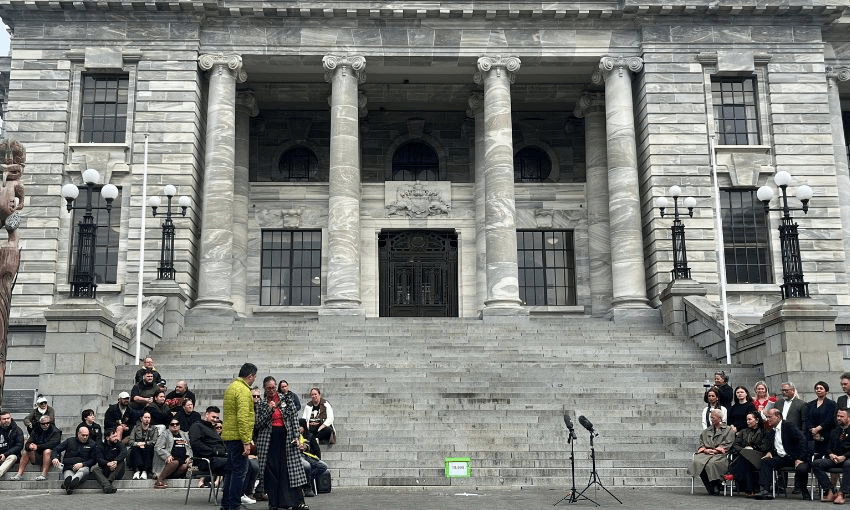 Two crowds sit outside the parliament building, in front of the parliament steps, with a petition box between them.