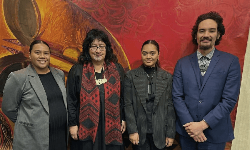 Four people stand side by side in front of a vibrant red mural. Three women and one man are dressed formally in suits and traditional Māori pendants, smiling or looking at the camera.