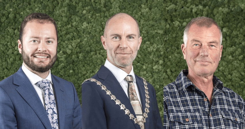 Three men stand in front of a green foliage background
