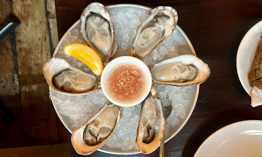 A plate of six raw oysters on the half shell arranged in a circle on ice, served with a lemon wedge and a cup of mignonette sauce in the center, on a dark table next to a partially visible plate.