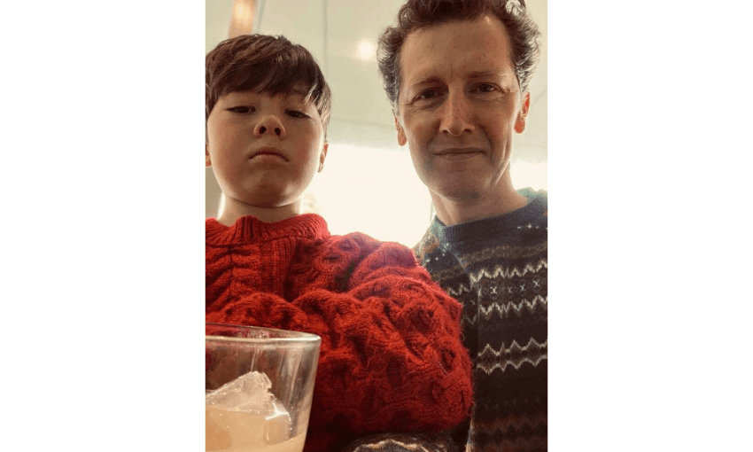 A photograph of a child wearing a red jumper and a man beside him. They are both looking into the camera lens. The child is serious. The man is smiling.