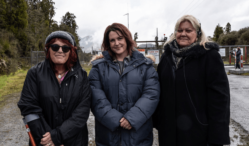 Three women stand on a road in Greymouth wearing big jackets and headphones. 