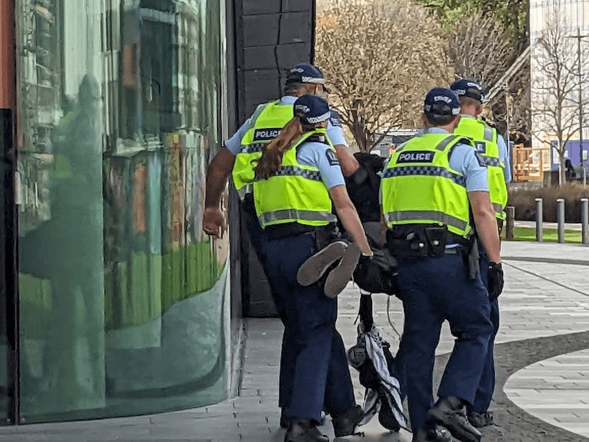 four hi-vis wearing police officers hold all four limbs of a protester with a kuffiya dangling from their hand