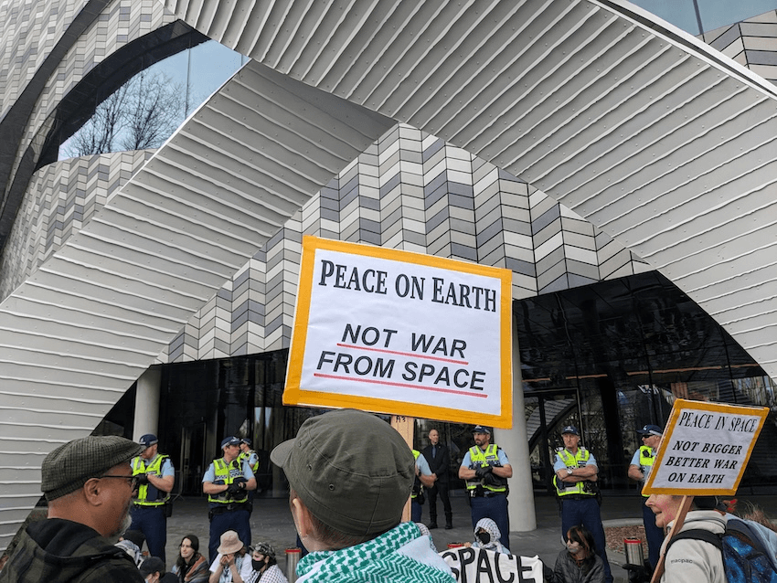 a sign saying peace on earth not war from space, with the grey tiles of Te Pae behind it