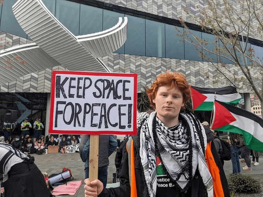 a red haired young man wearing a keffiyeh and holding a sign saying keep space for peace