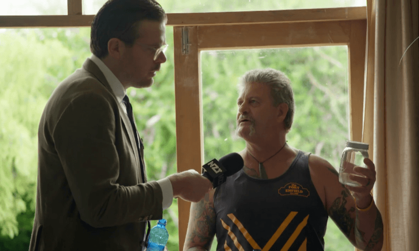 a tall man in a suit holds a microphone up to an older man in a construction singlet and a goatee