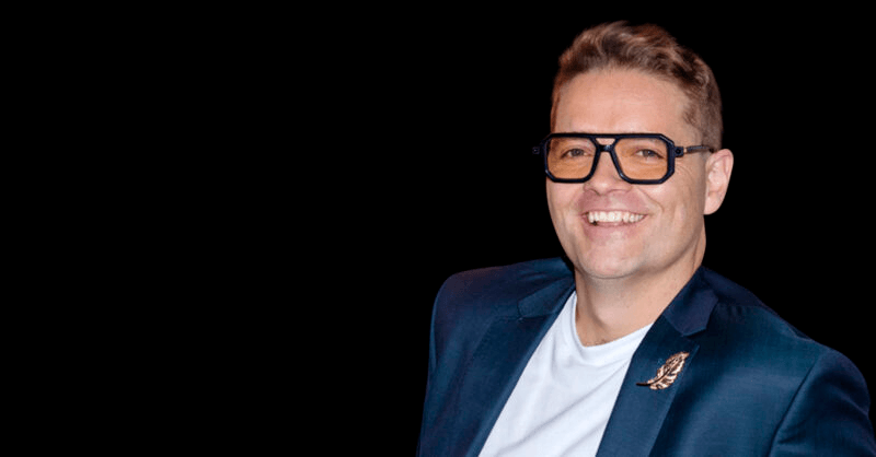 A man wears a white t-shirt and blue blazer and tinted glasses, smiling at the camera