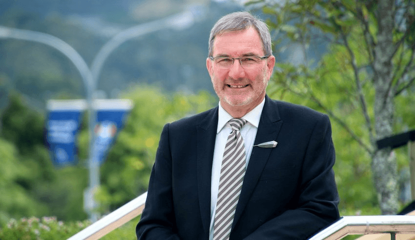 Paul Spoonley wears a suit and tie and smiles in front of some trees