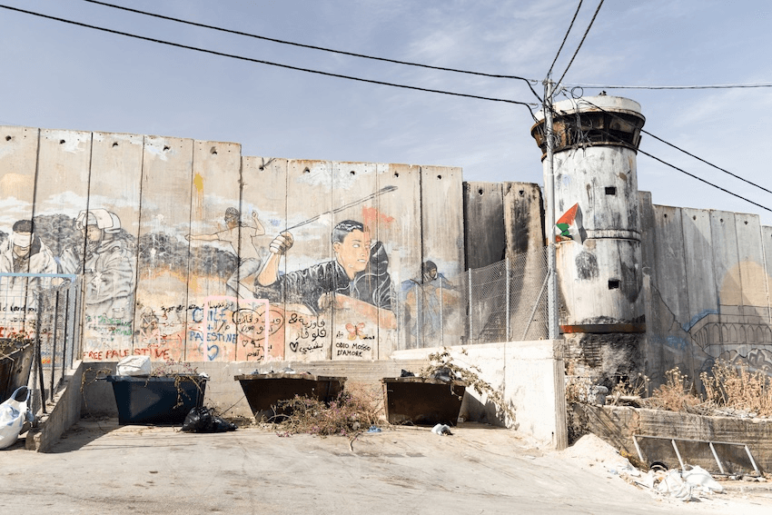 A concrete separation wall with political graffiti, including a figure slinging a stone. A worn watchtower stands to the right with a Palestinian flag. Trash bins and cables are visible in the foreground under a clear sky.