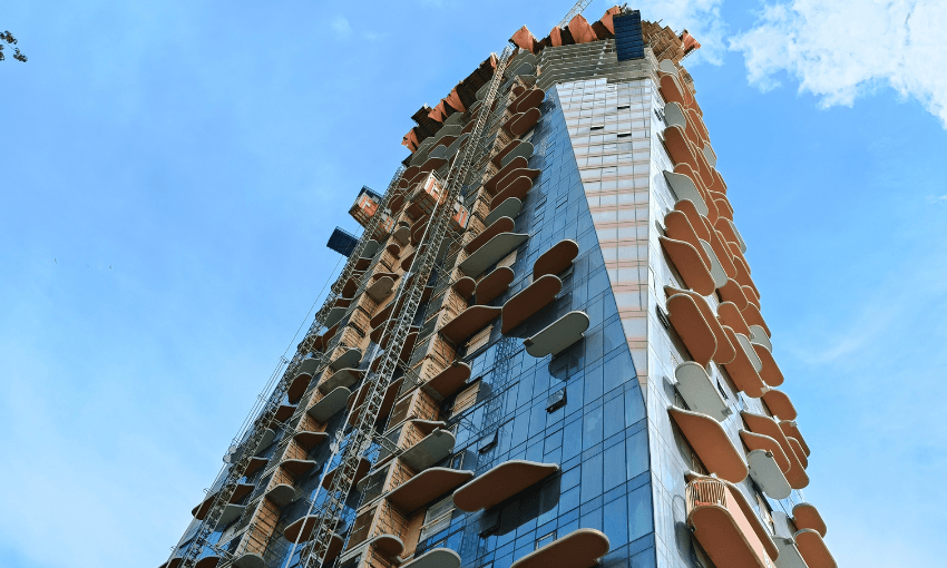A tall modern building under construction, featuring irregularly shaped balconies and glass panels, set against a bright blue sky with some clouds. Scaffolding and safety netting are visible along the structure.