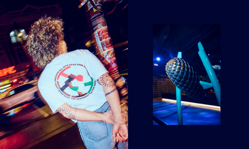 A person with curly hair stands on a city street at night, hands behind their back, wearing a white t-shirt with a colorful circular design. Beside them is a close-up of a black spiked microphone under blue lighting.