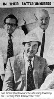 Three men pose indoors; the man in front wears a suit, tie, and a towelling hat, while the two men behind him wear glasses, shirts, and ties. A coat hook is visible on the wall behind them.