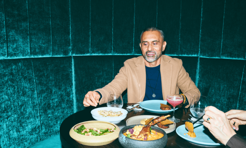 A man sits at a round table in a teal velvet booth, enjoying a meal with various dishes and drinks. He is smiling, and another person's hand is reaching for food across from him.